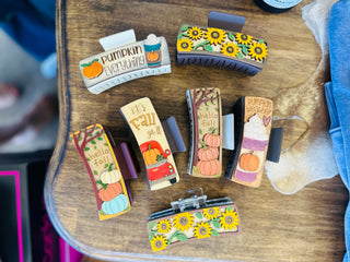 Seven handmade fall-themed hair claw clips featuring pumpkins, sunflowers, and autumn sayings displayed on wooden table.
