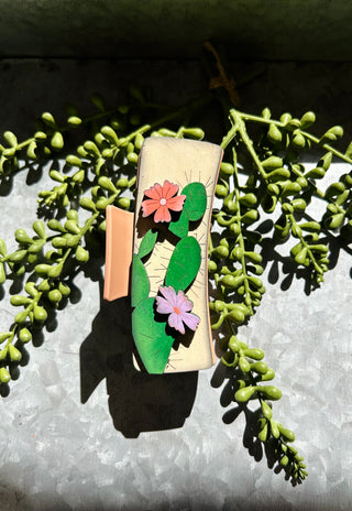Handmade wooden hair clip with painted floral design featuring green leaves and colorful flowers on succulent background.
