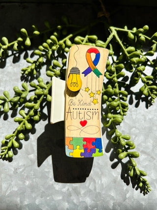 Handmade wooden bookmark with autism awareness ribbon design and "Be Kind" message displayed with succulent plants