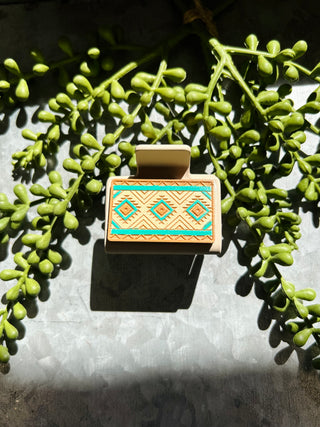 Handmade hair clip with geometric tribal pattern in turquoise and orange displayed among green succulent plants