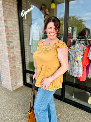 Woman wearing yellow embroidered floral top with tiered design, standing outside boutique storefront.