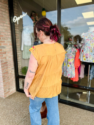 Woman wearing yellow embroidered floral top with tiered ruffle design, standing outside clothing store.