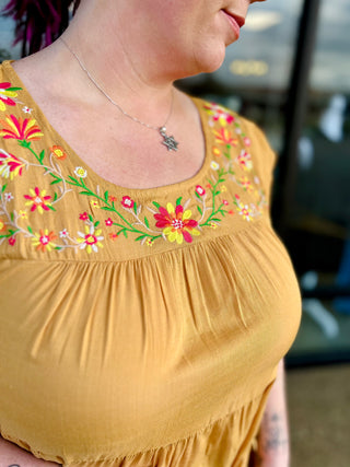 Woman wearing mustard yellow embroidered floral top with colorful flower details and gathered fabric design.
