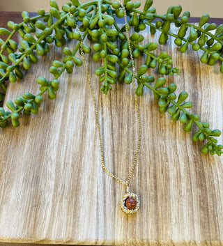 Gold chain necklace with circular pendant containing amber-colored stone, displayed on wood surface with green foliage.