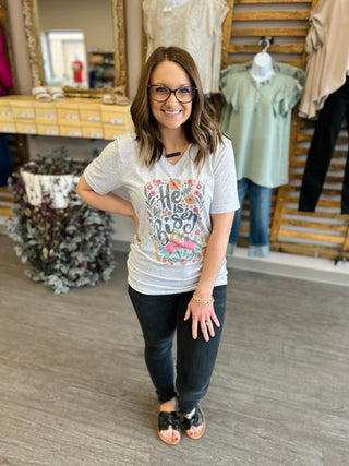 Woman wearing gray floral "He Is Risen" graphic tee in boutique clothing store setting