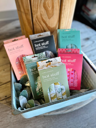 Collection of colorful heated neck wrap packages displayed in a metal container with decorative stones.