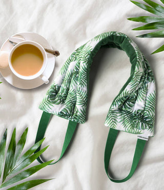 Heated neck wrap with tropical leaf pattern and green straps displayed with tea cup and palm leaves