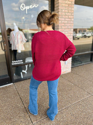 Woman wearing a long-sleeve waffle-knit sweater with high-low hem and blue jeans, back view.