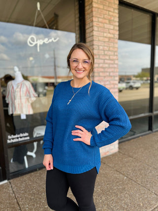 Woman wearing a relaxed waffle-knit sweater with long sleeves and curved high-low hem.
