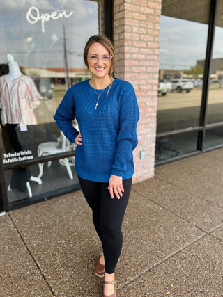 Woman wearing a long-sleeve waffle-knit round neck high-low sweater paired with black leggings.