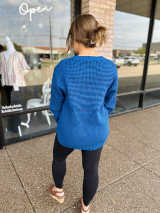 Woman wearing a waffle-knit round neck pullover sweater with high-low hem, back view.