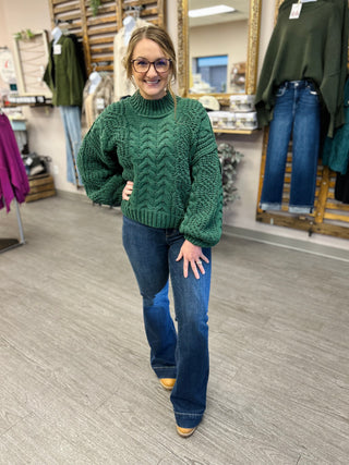 Woman wearing high-rise dark flare jeans with green cable-knit sweater in clothing boutique setting.