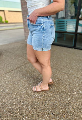 Woman wearing light wash denim high rise cuffed shorts with white top and pearl sandals outdoors