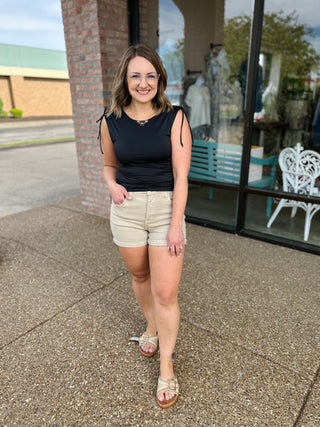 Woman wearing high-rise cuffed tan shorts with black sleeveless top and sandals, standing outdoors.