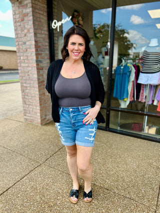Woman wearing high rise distressed cuffed denim shorts with black cardigan standing outside boutique storefront.