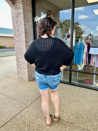 Woman wearing distressed denim cuffed shorts and black knit sweater standing outside boutique storefront