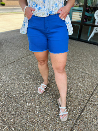 Woman wearing blue high-rise denim shorts with frayed hem and floral top, hands in pockets.