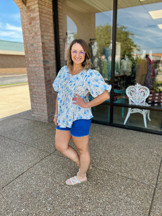 Woman wearing blue high-rise frayed shorts with floral top, standing outside storefront smiling.