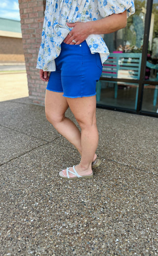 Woman wearing blue high-rise denim shorts with frayed hem and floral top, standing outdoors.
