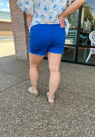 Woman wearing royal blue high-rise denim shorts with frayed hem and floral top, back view.