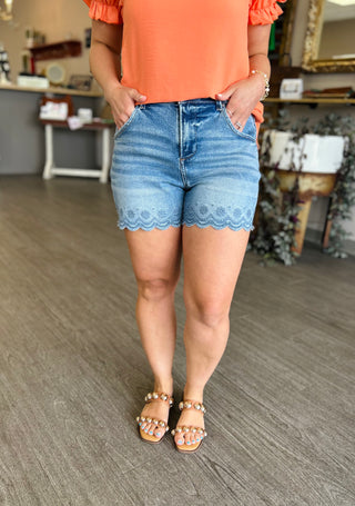Woman wearing high-rise denim shorts with scalloped hem detail and embroidered trim, paired with coral top.