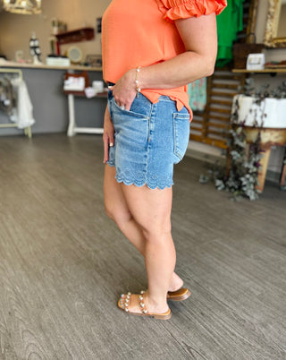 Woman wearing high rise denim shorts with scalloped hem and coral top in retail store setting