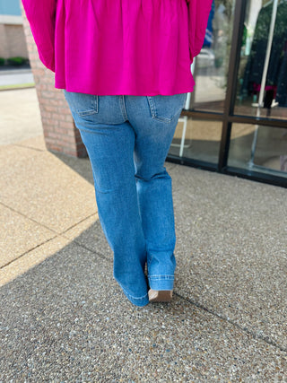 Person wearing high rise wide hem pintuck flare jeans with bright pink top, walking on concrete sidewalk.