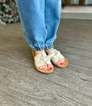 Person wearing blue denim joggers with elastic cuffs and beige bow-detail sandals on wooden floor.