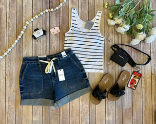 Casual summer outfit flatlay with denim shorts, striped tank top, sandals and accessories on wooden background