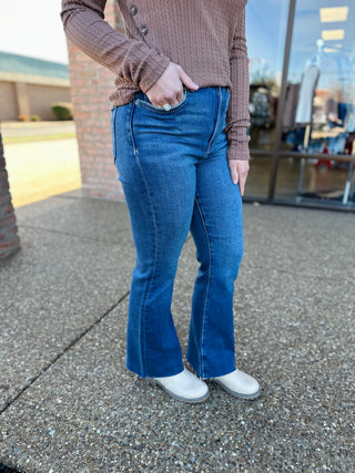 Person wearing dark wash cropped flare jeans with raw hem and brown knit sweater standing outdoors.