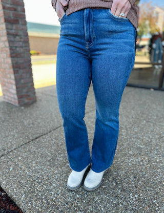 Person wearing high-rise dark wash cropped flare jeans with raw hem and white boots on pavement.