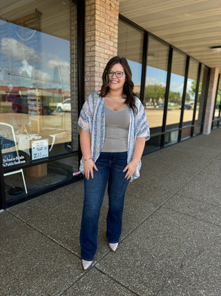 Woman wearing dark wash skinny bootcut jeans with gray top and patterned kimono outside storefront.