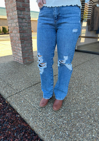 Person wearing medium wash distressed denim jeans with ripped knees and frayed details, standing on concrete sidewalk.