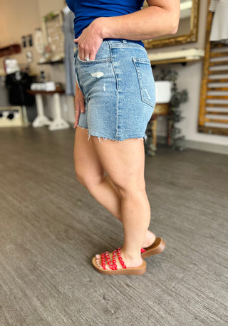 Woman wearing distressed denim shorts with raw hem and frayed details, paired with red studded sandals.