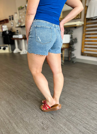 Woman wearing distressed denim shorts with raw hem and blue top, viewed from behind in kitchen setting.