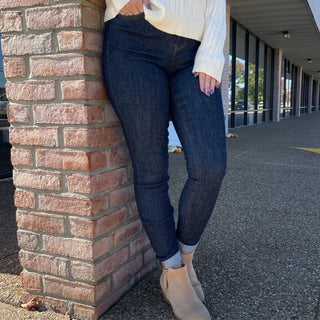 Woman wearing dark wash skinny jeans with embroidered pockets, white sweater, and tan ankle boots against brick wall.