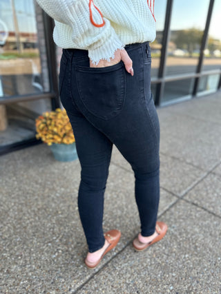 Woman wearing high-waisted black skinny jeans with white sweater and brown slides on concrete sidewalk.