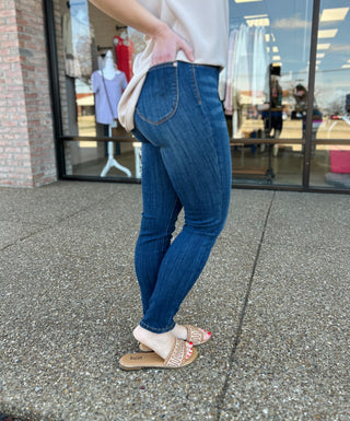 Woman wearing dark wash high-waisted pull-on skinny jeans with white top and sandals outside boutique.