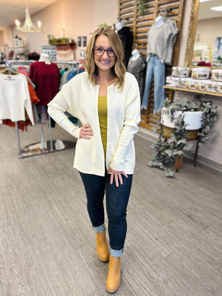 Woman wearing ivory open-front cardigan over yellow top with jeans and boots in clothing boutique