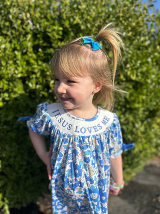 Young girl wearing blue floral "Jesus Loves Me" dress with white collar outdoors in garden setting.