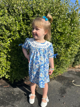 Young girl wearing blue floral "Jesus Loves Me" dress with puffed sleeves standing outdoors