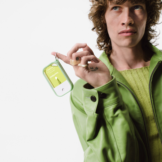 Person in green jacket holding a Touchland Hand Mist Case keychain accessory on finger.