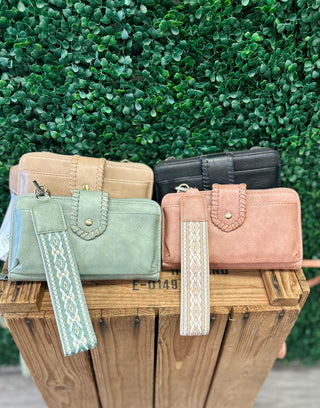 Four crossbody wallets with decorative wrist straps displayed on wooden crate against green foliage background.