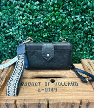Black leather crossbody wallet with patterned wrist strap displayed on wooden crate against green foliage background.