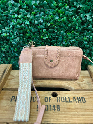 Pink leather crossbody wallet with decorative stitching and patterned wrist strap on wooden surface with greenery background.