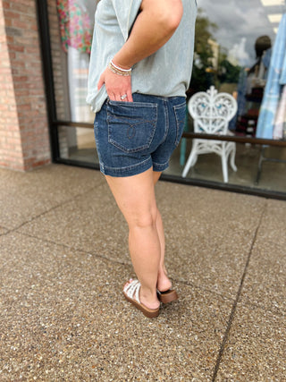 Woman wearing Judy Blue high-waisted denim shorts with white tank top and sandals outdoors.