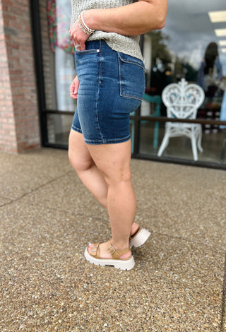 Woman wearing high-waisted denim bermuda shorts with pockets, styled with sandals and casual top.
