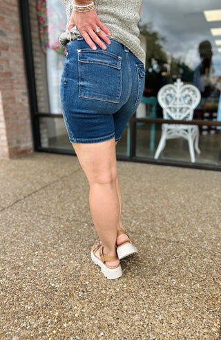 Woman wearing high-waisted denim bermuda shorts with back pockets, styled with casual top and platform sandals.