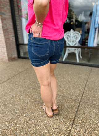 Woman wearing high-waisted denim shorts with pink top and sandals, showing back view and fit.