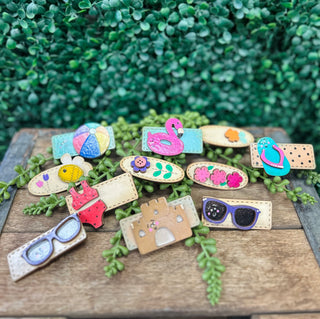 Colorful handmade kids hair clips with fun designs including sunglasses, flamingo, flowers, and beach themes displayed out...